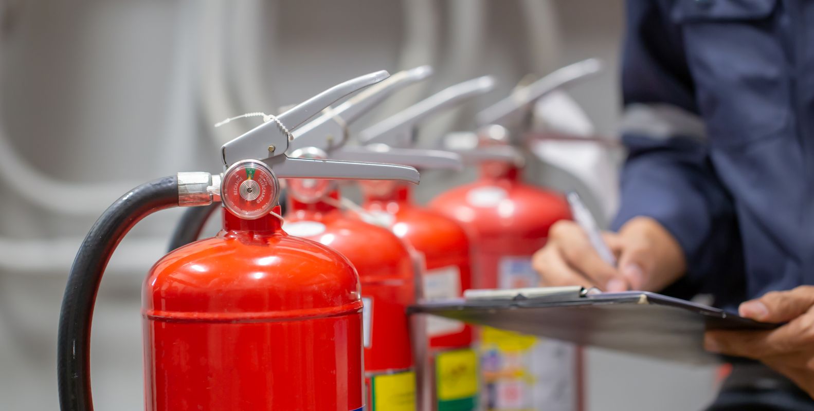 Mehrere rote Feuerlöscher stehen nebeneinander, eine Person prüft sie mit Klemmbrett